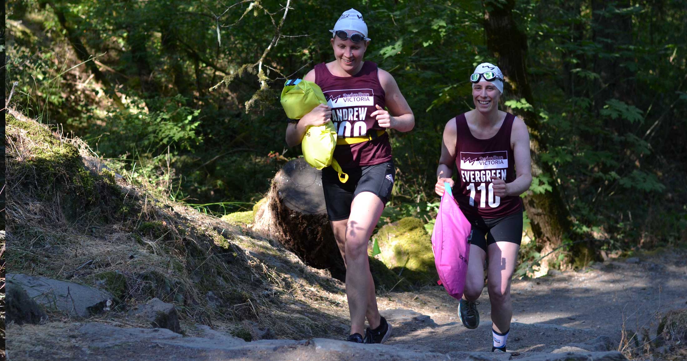 Two athletes race together, smiling, through the forest trail on their way to the next swim leg.